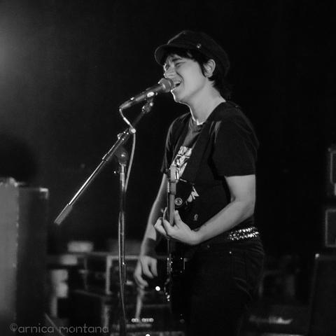 A performer stands on stage, playing an electric guitar and singing into a microphone under bright lights. The image is in black and white, capturing the energy of a live music performance. The performer wears a graphic t-shirt and a hat, and is expressive