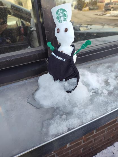Tiny  barista snowman wearing a Starbucks mask for an apron, with coffee stirrer arms, coffee bean eyes, and a Starbucks logo sticker hat, perched on the drive thru window ledge.