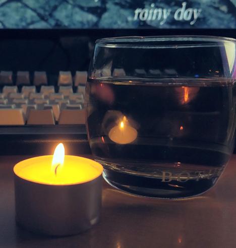 **TL;DR:** A quiet, reflective moment with a candle, a glass of drink, and a melancholic caption about an annual tradition, loneliness, and a heartfelt "kampai" (cheers).  

The image shows a small, lit tealight candle next to a glass of liquid, with the candle's warm glow reflected in the glass. In the background, a keyboard and a screen display the words "rainy day," adding to the somber, introspective mood. The caption references a familiar, possibly lonely ritual, with the phrase "Same procedure as every year?"—a nod to the classic sketch *Dinner for One*. The hashtags and broken-heart emoji convey a sense of loneliness, with "kampai" (cheers) suggesting a solitary memorial toast.