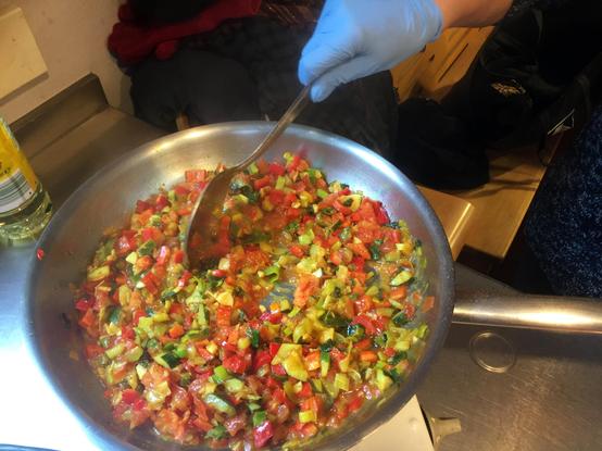 Eine Besucherin von Retten Teilen Helfen kocht in einer Bratpfanne klein geschnittenes Gemüse. Sie rührt mit einem großen Löffel. Der Gemüse-Mix besteht u.a. aus Zucchini, Porree und Paprika und wird für den Frühstückstisch vorbereitet. Alle Lebensmittel sind gerettet.