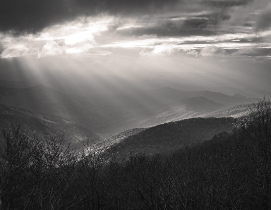 Sun rays distinctly burst through the clouds over a blue ridge mountain landscape, highlighting the valley below 