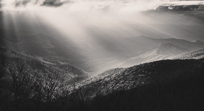 Sun rays distinctly burst through the clouds over a blue ridge mountain landscape, highlighting a ridge in the foreground and a valley below 