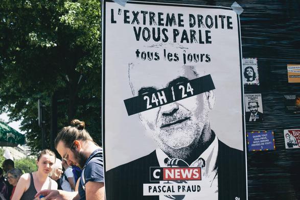 Photographie d'une affiche prise au cours d'une manifestation. Sur cette dernière, nous pouvons lire "l'extrême droite vous parle tous les jours 24H / 24, Cnews, Pascal Praud" avec le portrait de l'animateur vedette de la chaîne les yeux masqués par le texte "24H / 24".
