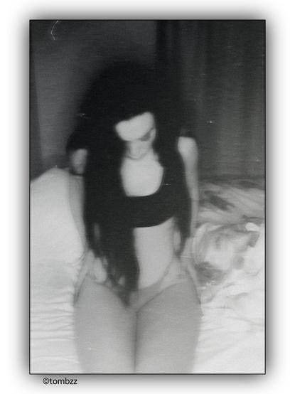 A young woman is sitting on a bed. Her head is bowed. She is wearing a black top and underwear. Her long hair falls over her shoulders and down her front. Her posture is slightly slouched, with her, conveying a sense of resignation and emotional heaviness. The background includes a curtain and some bedding. The image has a grainy, black-and-white effect, giving it a vintage or artistic feel.