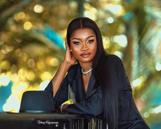 A pretty female model with dark hair and black dress looks straight into the camera. Her black hat rests on the table beside her, which she is leaning onto. The palm trees in the background are blurred.