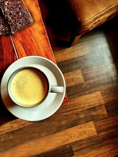 A photo of a grey cup of coffee which is standing at the edge of a brown table. Will it drop down?On the right side of the image you see a brown chair. On the table are 2 slices of chocolate cake.