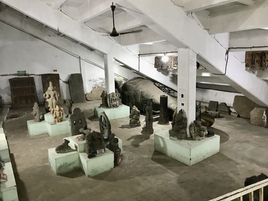 Das Foto zeigt einen Blick in den Ausstellungsraum des Government Museums unter den Sitzplätzen des Nehru Stadium in Coimbatore. Zu sehen sind zahlreiche Steinskulpturen und Steintafeln mit Inschriften, die teils um die Stützpfeiler herum arrangiert wurden. Die Artefakte stammen aus einer Zeit zwischen dem 12. bis 19. Jahrhundert.