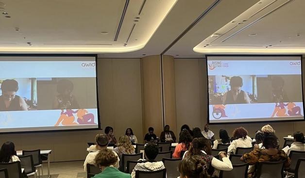 A conference room with attendees seated and listening to a panel discussion. The panelists, a diverse group of speakers, are seated at the front, while two remote participants appear on large screens displaying the "AWID" and "Rising Together" forum logos. The event focuses on transnational solidarity and addressing technology-facilitated gender-based violence (TFGBV).