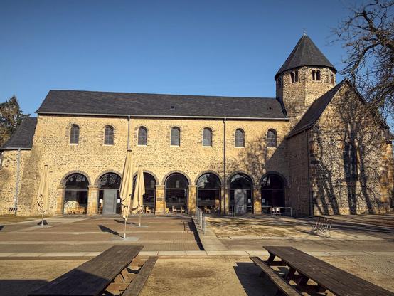 Das Foto zeigt das Kloster Schiffenberg in Gießen an einem sonnigen Februartag. Die romanische Kirche mit ihrem markanten Rundturm und den Rundbogenfenstern dominiert das Bild. Die Fassade aus hellem Sandstein ist von klaren Linien geprägt, und die Arkaden im Erdgeschoss deuten auf eine gastronomische Nutzung des Gebäudes hin. Vor dem Kloster befinden sich mehrere geschlossene Sonnenschirme sowie Holzbänke und Tische, die einen einladenden Platz zum Verweilen bieten. Die langen Schatten der Bäume auf dem gepflasterten Platz und der tiefblaue Himmel verstärken die ruhige und historische Atmosphäre des Ortes.