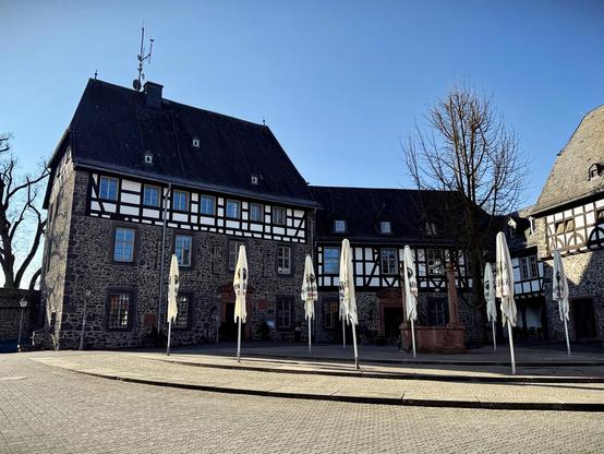 Das Foto zeigt das Kloster Schiffenberg in Gießen an einem sonnigen Tag. Im Vordergrund stehen geschlossene Sonnenschirme auf einem gepflasterten Platz. Die historischen Fachwerk- und Bruchsteingebäude mit dunklen Schieferdächern prägen das Bild. Ein blattloser Baum steht vor der Fassade, der Himmel ist klar und blau.
