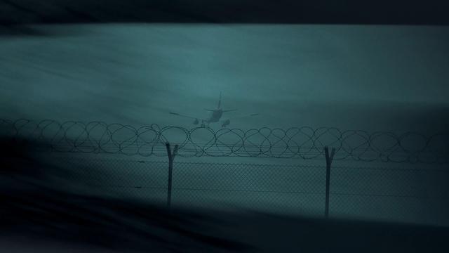 Viewed from the back, a plane is about to land on an airstrip. Between the plane and the camera, there's a fence with barbwire on top, the plane is eemingly touching it with its landing gear. The whole photo is tinted in greyish-green and has a vignette-effect around it, as if the camera was looking through a peephole.
