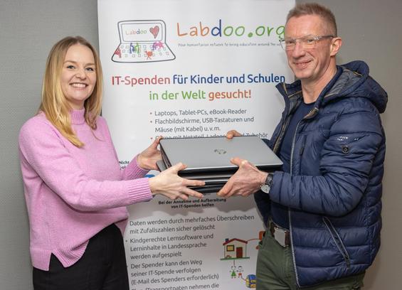 Vor einem Rollup von Labdoo steht links Leandra von Labdoo und rechts Marko von eGo MV. Zwischen sich halten beide einen Stapel gespendeter Laptops. Beide schauen in die Kamera.