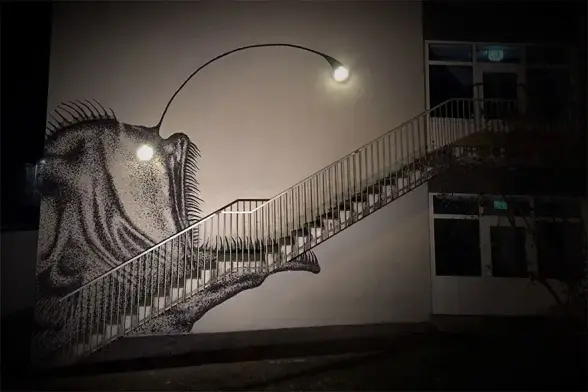 Mural of an anglerfish that is painted adjacent to a flight of stairs. Lightbulbs on the wall coincide with the fish's eye and it's dangling bits used to lure prey. It creates a cool effect in darkness with the lights on and in daylight as well. Great work by artist Skurk and shared originally by @streetartutopia