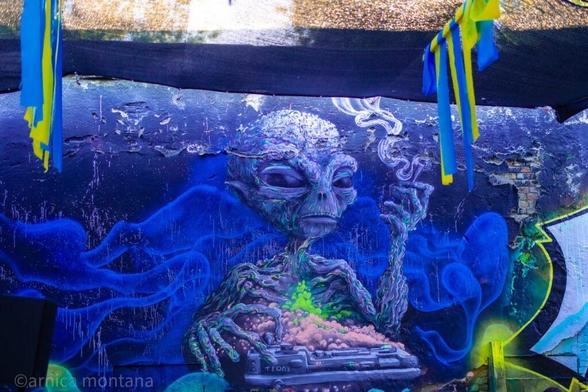 A vibrant mural depicting an alien figure sitting with a computer, smoking and surrounded by colorful swirling patterns. The background features shades of blue and green, with some peeling paint adding texture. Colorful ribbons hang from the top, enhancing the artistic atmosphere.