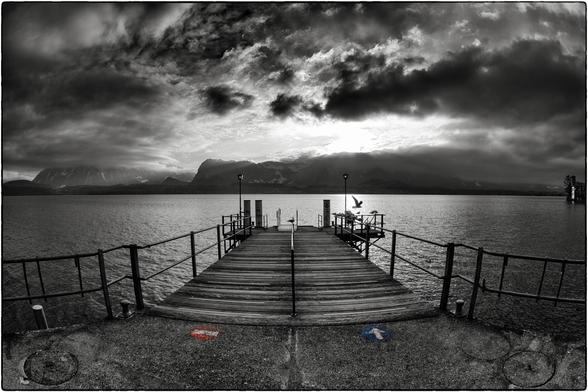 Schwarzweissfotografie mit Selektivfarben des Landungsstegs in Oberhofen am Thunersee unter dramatischem Gewölk. 