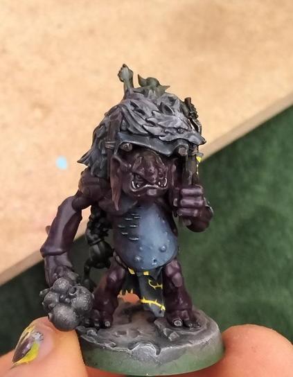 Picture showcasing a warhammer troll miniature, Uglug, from the warhammer underworlds warband "Borgritt's beastrabbaz" , a rock troll with some fungus growing on their right side of the body, with an "unibrow" that's just their nose ridge extending from the top of the head to the regular point where it grows, and clutching a wolf pelt skin over them. A goblin is sitting atop the troll. A casket with a hanging goblin is hanged at the back of the troll as well.The loincloth and cloth scraps are painted in black with yellow "spikes and spiky roots" patterns