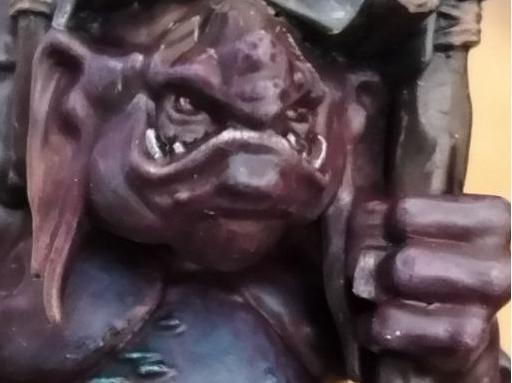 Picture showcasing a warhammer troll miniature, Uglug, from the warhammer underworlds warband "Borgritt's beastrabbaz" , a rock troll with some fungus growing on their right side of the body, with an "unibrow" that's just their nose ridge extending from the top of the head to the regular point where it grows, and clutching a wolf pelt skin over them. A goblin is sitting atop the troll. A casket with a hanging goblin is hanged at the back of the troll as well.The loincloth and cloth scraps are painted in black with yellow "spikes and spiky roots" patterns. Closeup of the face
