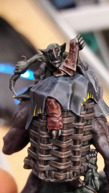 Picture showcasing a warhammer troll miniature, Uglug, from the warhammer underworlds warband "Borgritt's beastrabbaz" , a rock troll with some fungus growing on their right side of the body, with an "unibrow" that's just their nose ridge extending from the top of the head to the regular point where it grows, and clutching a wolf pelt skin over them. A goblin is sitting atop the troll. A casket with a hanging goblin is hanged at the back of the troll as well.The loincloth and cloth scraps are painted in black with yellow "spikes and spiky roots" patterns.closeup of the gobling at the top of the basket on the troll's back.