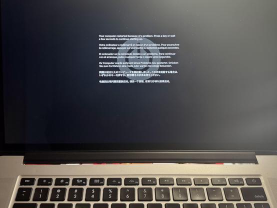 A Mac laptop screen displaying a kernel panic error message in multiple languages. The message states that the computer restarted due to a problem and prompts the user to press a key or wait a few seconds to continue starting up. The background is black, and the keyboard is visible.