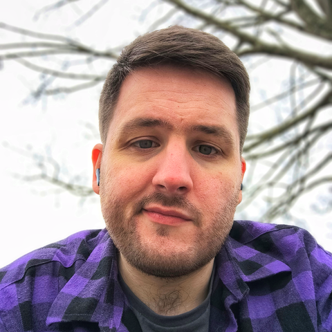 Selfie of a 30 something year old white man sitting in a park under a tree wearing a purple plaid shirt and a gray t-shirt, he has a light stubble on his face and a fresh haircut (short back and sides)