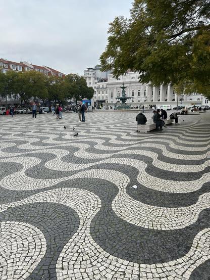 A bustling public square with a patterned cobblestone pavement featuring wavy designs. There are people walking, sitting on benches, and some pigeons scattered around. In the background, buildings and trees are visible, along with a fountain. 