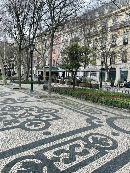 A decorative cobblestone walkway with intricate patterns leads through a park-like area featuring bare trees and colorful flower beds. Historic buildings in muted colors are visible in the background, along with outdoor seating and a lamppost.