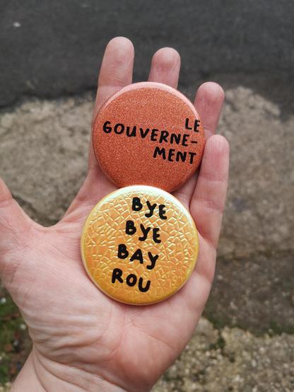Main tendue sur laquelle deux badges sont posés, avec écrit dessus "le gouverne-ment" et "bye-bye Bayrou"