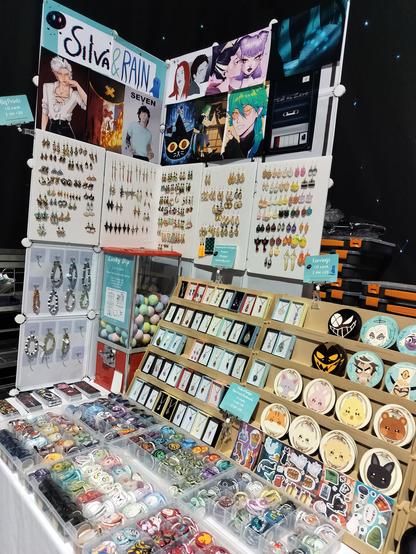 A picture of a double table set-up at Leeds Comic-Con. This is a close-up of the left side of the tables where you can see prints, earrings, bracelets, necklaces, coasters, badges and sticker sheets. You can see merch of different fandoms like Hermitcraft, Hazbin Hotel, Ghibli, Don't Starve, QSMP, Vocaloid, League of Legends, Generation Loss, ATEEZ, Tower of God and more.
