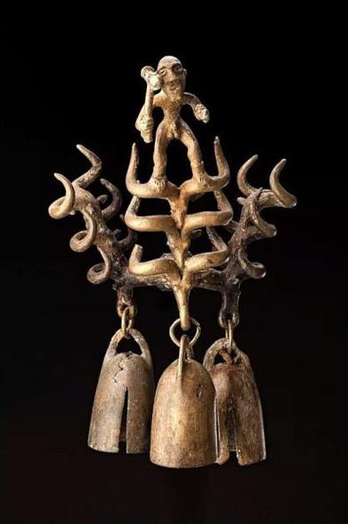 Artefacto metálico con tres columnas de tres cabezas cada una de uros. Cada cabeza inferior sostiene una campana, tres en total.  Sobre una de las cabeza superiores, hay una figura masculina desnuda, salvo por las botas, con un martillo en la mano derecha.