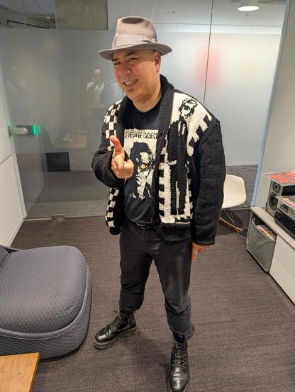 A man is posing indoors, smiling and pointing toward the camera with his right hand. He is wearing a gray fedora, a black t-shirt with a graphic design, black pants, and a hand-knitted black-and-white patterned cardigan featuring a ska or two-tone style design. The cardigan has an image of Walt Jabsco, the iconic dancing man associated with 2-Tone Records. He also wears black boots. The background includes a modern office setting with glass walls, a chair, a stereo system, and some furniture.