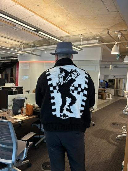 The same man is shown from behind, standing in an open-plan office with desks, computers, and modern lighting. His black-and-white cardigan prominently displays a large knitted graphic of Walt Jabsco, the iconic dancing ska figure from 2-Tone Records, framed by a checkered pattern. He is also wearing the gray fedora and black pants. The office has an industrial-modern style with exposed ceiling elements and a mix of furniture.