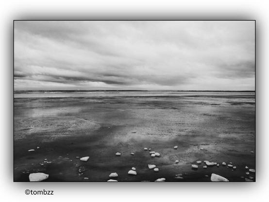 A black and white photograph of a frozen bay. The surface of the bay is covered with ice, and scattered pieces of ice are visible on the frozen surface. The sky above is filled with thick, overcast clouds, creating a moody and dramatic atmosphere. The horizon is barely visible in the distance, blending with the cloudy sky.