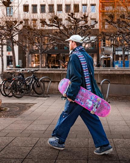 Ein junger Mann in sportlicher Kleidung geht über einen gepflasterten Gehweg in einer Stadt. Er trägt eine weite dunkelblaue Hose, ein passendes weites Oberteil und blaue Sneaker mit dicker Sohle. Um den Hals hat er einen bunten gestreiften Schal gelegt, und auf dem Kopf trägt er eine beigefarbene Baseballkappe. Um seinen Hals hängen große Kopfhörer. In der rechten Hand hält er ein pink-lila Skateboard mit auffälligem Muster und abgenutzten Rollen. Seine Haltung ist entspannt, während er nach vorne geht und dabei in sein Smartphone schaut. Im Hintergrund sind kahle, beschnittene Bäume, Fahrräder, ein moderner Gebäudekomplex mit großen Fenstern und eine Baustelle mit einem Gerüst zu sehen. Die Szenerie wirkt urban und modern, mit einer Mischung aus Architektur, Natur und städtischer Infrastruktur.