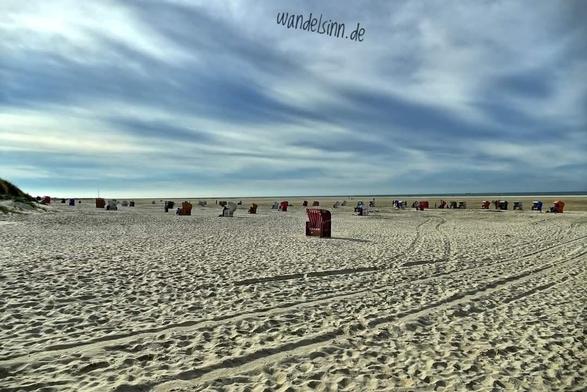Ein weißblauer Himmel, wie gemalt sieht er aus. Ein weiter Strand ist zu sehen,  das Wasser kann man fast nur ahnen. Einen Strandkorb kann man ganz gut erkennen. Viele weitere Körbe sind in Richtung der Wasserkante zu ahnen. Im Sand sind Fahrspuren und Fußabdrücke zu erkennen. Links die Ausläufer einer Düne.