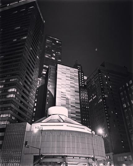 Black and white photo of a disc-shaped concrete building sitting at the intersection of Wabash and Wacker, along the Chicago River. It is nighttime, and various skyscrapers loom in the background. 
