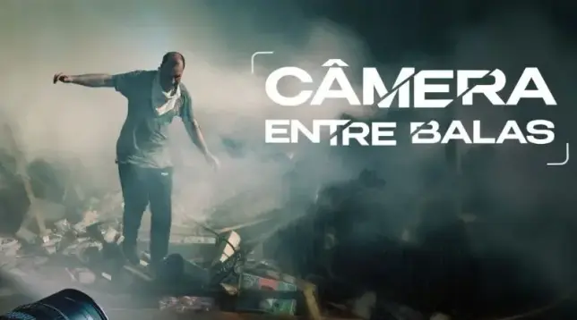 Pôster promocional da série documental "Câmera Entre Balas". Mostra um local aparentemente destruído por um bombardeio, esfumaçado, em meio ao qual um indivíduo se movimenta tentando se equilibrar entre os escombros.