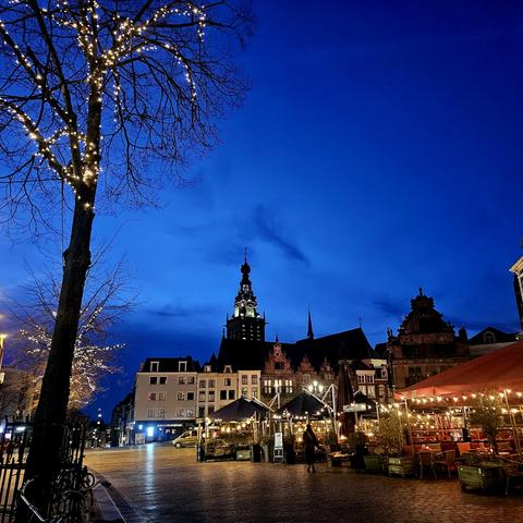 The image shows the Grote Markt in Nijmegen during twilight. The sky is a deep blue, and the historic buildings, including the Waag and the Stevenskerk, are beautifully illuminated. In the foreground, trees adorned with festive lights and a cozy terrace with lit tents and seating create a warm atmosphere. The cobblestone street adds to the charm, with a few people visible in the illuminated cityscape.