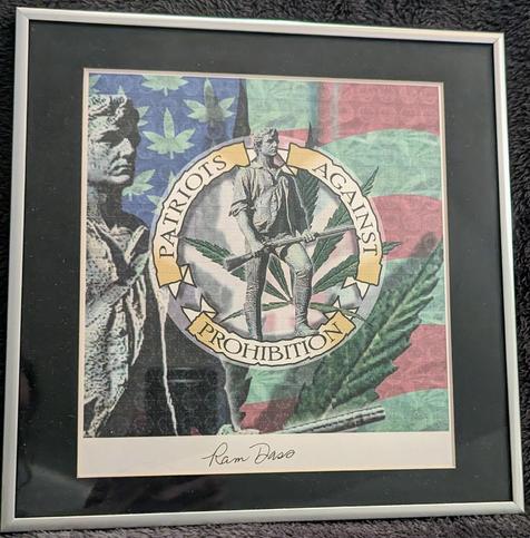 The image is a framed LSD Blotter Art print signed by BaBa Ram Dass. In the center of the frame, there is a circular emblem with the words "Patriots Against Prohibition" written in gold lettering. The emblem is surrounded by a green marijuana leaf and an American flag. On the left side of the emblem, there are two figures of a minuteman holding a rifle in his hands ready to defend your freedom standing in front of the flag background. The image is framed in a silver frame.