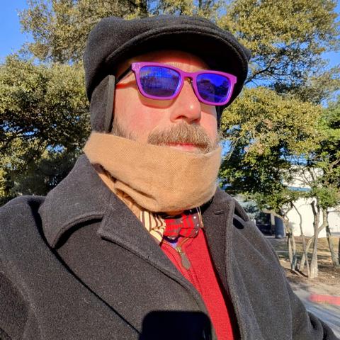 A photo of me, a 46 year old white man with a shaved head, a short cropped brown & grey beard, 

photographed from the chest up

In front of some live oak trees in an office park,

wearing

rectangular purple acetate glasses, a red and cream Ankara style bowtie on a purple dress shirt under a deep red quarter zip sweater with a black topcoat and black flat cap with ear flaps. A camel colored scarf hangs around my neck.
