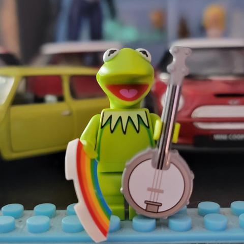 Close up picture of Lego minifigure Kermit the Frog holding a banjo in his left hand and a rainbow in his other hand with a picture of some cars out of focus behind him.