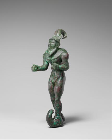 Figura humana de aleación de cuernos. Tiene ambos brazos paralelos, con los codos en ángulo recto y un pie adelantado. Tiene barba, zapatos con punta larga y curva. Tiene un gorro con orejas y dos cuernos curvos y pegados. Sobre los hombros cuelgan dos alas que se unen en el pecho.
