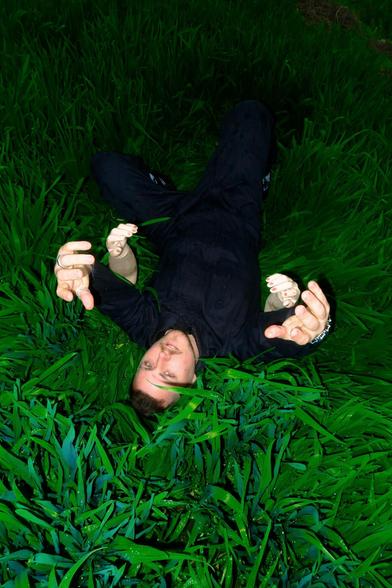 Nuño de la Serna lying on the ground amidst tall green grass, reaching upwards with both hands, during a photoshoot in Sichuan, China