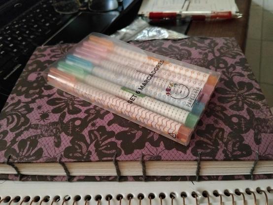 Package of 6 pens that provide different patterns when rolled on paper sits on top of a black and purple journal. Miscellaneous office supplies are strewn around in the background.