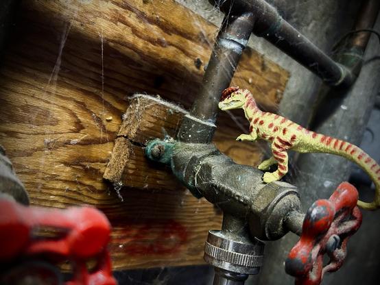 A green Dilophosaurus with brown spots stands on a tap with a red handle and covered in cobwebs. A piece of wood is behind the taps and looks worn and damaged. A second red faucet handle is in the foreground and partially blurred.