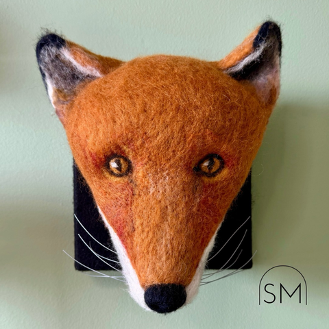 Felted fox head mounted on a black, felt covered canvas. Face colours are shades of amber, white, and cream. Black nose and grey, white, and black ears. Whiskers are made of beading wire.