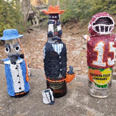Thee Decorated bottles stand next to each other. The first one is decorated to look like a fisherman, the second like a cowboy, & the third like a football quarterback.