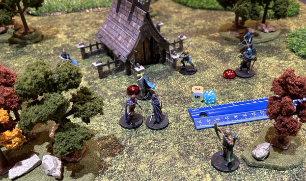 A photo of a tabletop wargame in progress. A forested area around a mausoleum is the setting of a skirmish between some blue-uniformed enemies and a ragtag group of fighters. All are soldiers with bows, swords, polearms, etc.