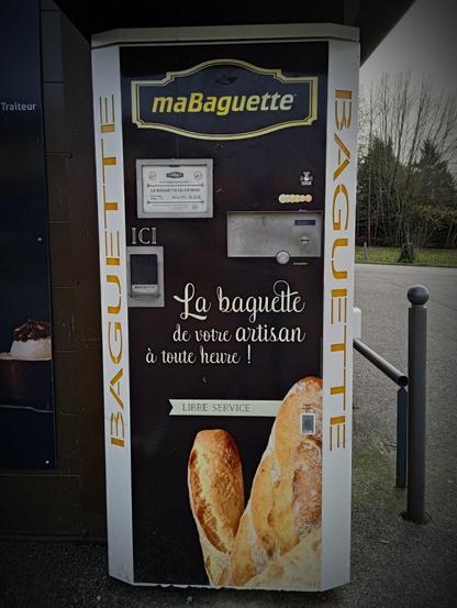 A vending machine standing in a carpark by a bakery door. The word BAGUETTE is written vertically in orange letters down the sides, a large picture of baked bread on the front. "Baguette, from your artisan, at any hour!" is written in French, in curly whitescrupt across the front. There is a money slot, and a little door reading ICI (HERE) That presumably dispenses one's freshly baked bread