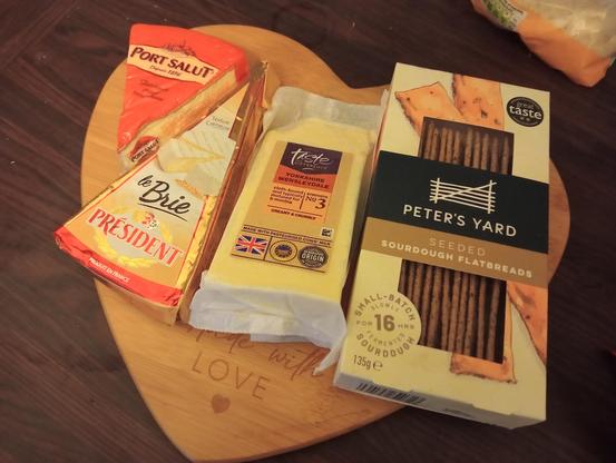 A heart-shaped wooden board with 3 types of cheese, and fancy crackers on top (Port Salut, French Brie and Yorkshire Wensleydale)