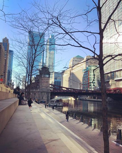 View of the Merchandise Mart from the steps of the Chicago Riverwalk. Pedestrians are walking along the sidewalk and others are sitting on the steps near me (the photographer). 